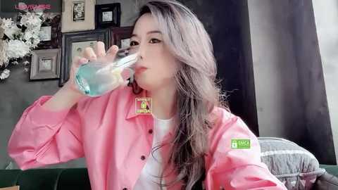 Media: Video of an Asian woman with long black hair, wearing a pink button-up shirt, drinking from a clear plastic cup, in a cozy room with framed photos and a green couch.