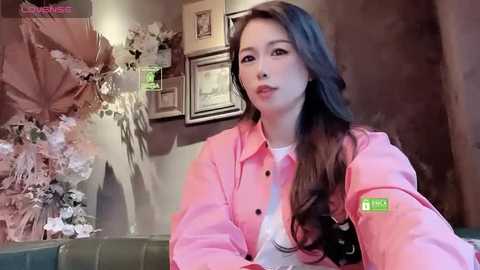 Media: A video of a young Asian woman with long brown hair, wearing a pink denim jacket over a white shirt, sitting in a cozy living room with framed artwork and floral decorations in the background.