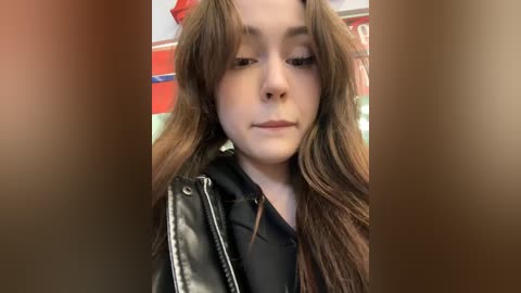 Media: Video of a young Caucasian woman with long, wavy brown hair, wearing a black leather jacket and a black shirt. The background is blurred, featuring red and white colors.