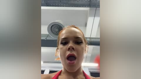 Media: A video of a woman with light skin and brown hair, wearing a red top, mid-sentence with mouth open, in an airplane cabin with a vent and blurred background.