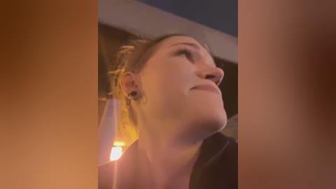 Media: Video of a Caucasian woman with light skin, dark hair tied back, looking upward with a serene expression. Warm, soft lighting casts a golden glow, creating a cozy ambiance.