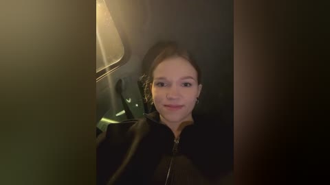 Media: A video of a young woman with fair skin and brown hair, smiling, sitting in a dimly lit car interior with a beige lamp and window visible.