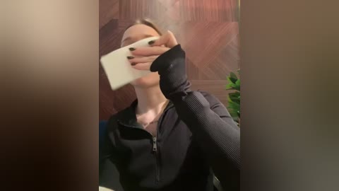 Media: Video of a woman with fair skin, wearing a black zip-up jacket, holding a white phone to her face, partially obscured by a blurred foreground. Background features a wooden wall and a green plant.