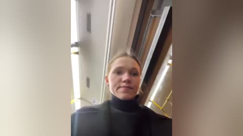 Media: A video of a young, fair-skinned woman with light brown hair, wearing a black turtleneck sweater, standing in a cluttered, dimly lit room with exposed pipes and a white ceiling.