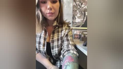 Media: Video of a young woman with light skin and shoulder-length brown hair, wearing a plaid shirt and black tank top, sitting in a kitchen with a floral-patterned curtain and various objects on the counter.