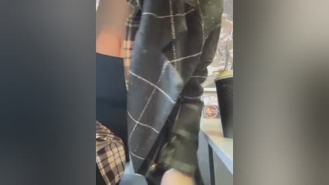 Media: Video of a person wearing a black and white plaid jacket with white stitching, sitting at a table. Background includes a cup, a phone, and a patterned chair.