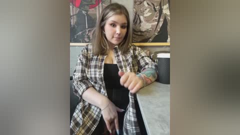 Media: Video of a young woman with light skin, shoulder-length brown hair, and a black top, wearing a plaid shirt, gesturing with her right hand. She has tattoos on her left arm. Background includes abstract art and a countertop.