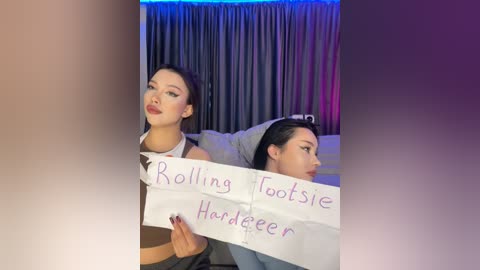 Media: Video of two young women with light skin, wearing makeup, holding a sign reading \"Rolling Footsie Harder\" in a dimly lit room with gray curtains and blue lighting.