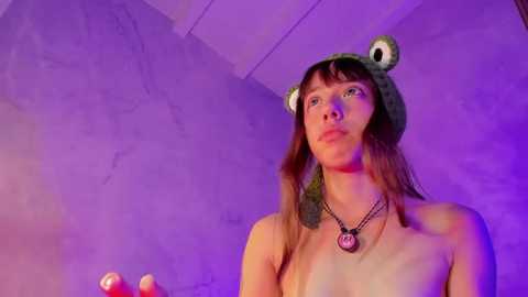 Media: Video of a topless, fair-skinned woman with long brown hair, wearing a frog hat and a pink necklace, against a purple-lit, concrete wall. She appears thoughtful.