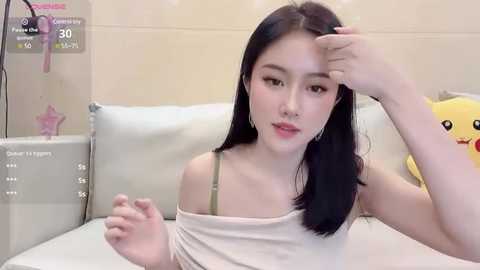 Media: Video of an East Asian woman with long black hair, wearing a beige top, sitting on a white couch. Background includes a plush yellow duck toy and a star-shaped ornament.