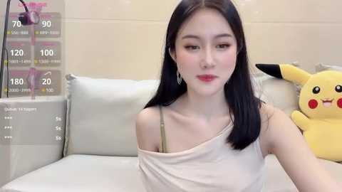 Media: Video of a young Asian woman with long black hair, wearing a white spaghetti strap top, sitting on a white couch. Pikachu plush toy is behind her.