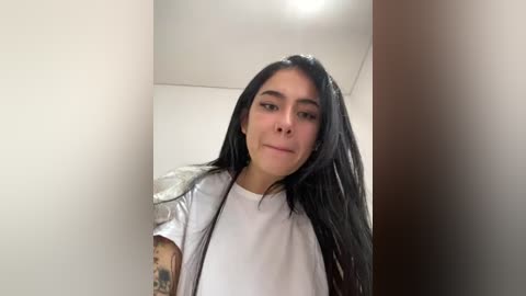 Media: Video of a young woman with long, straight black hair, light skin, and a medium build, wearing a white T-shirt with a colorful tattoo sleeve, standing indoors with a plain white wall and a wooden door in the background.