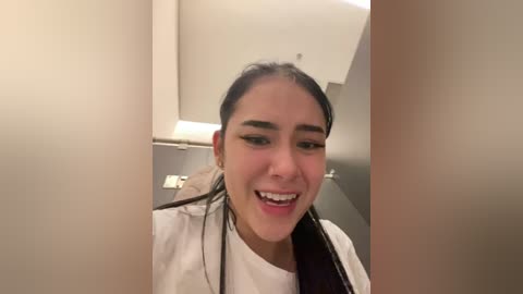 Media: Video of a young woman with long dark hair, light skin, and a cheerful expression. She's wearing a white jacket and black shirt, partially obscured by beige curtains. Background shows a modern, minimalist bathroom with beige walls and a white ceiling.