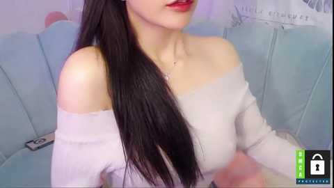 Media: Video of an East Asian woman with long black hair, wearing a white off-shoulder top, sitting on a light blue couch in a softly lit room.