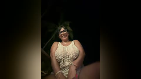 Media: Video of a smiling, curvy woman with glasses, wearing a white, lace-up top, seated in a dark, dimly-lit room.