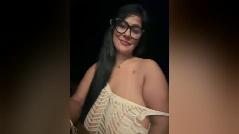 Media: Video of a smiling, light-skinned woman with long black hair, wearing large glasses and a white, knitted, off-shoulder top, against a dark background.