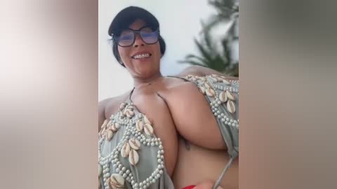 Media: A video of a smiling, short-haired Asian woman in a revealing, embellished gray bikini with pearl details, wearing large black-framed glasses. Background includes a blurred palm tree.