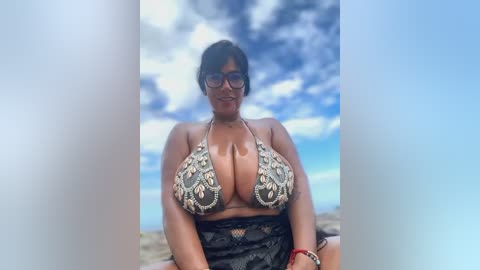 Media: Video of a curvaceous woman with medium-dark skin, large breasts, and glasses, wearing a black lace skirt and a detailed, multi-colored bikini top. She sits on a sandy beach with a clear blue sky and scattered clouds in the background.