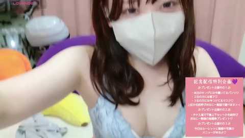 Media: Video of an East Asian woman with long, dark hair, wearing a white mask and a light blue lace bra, taking a selfie. Background includes a purple chair and a yellow object. Text overlay in Japanese.