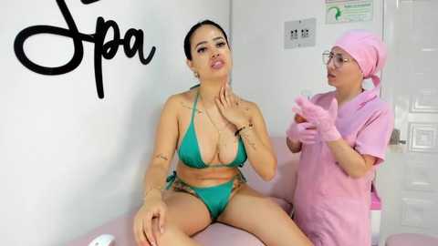 Media: Video of a Latina woman in a green bikini with large breasts and a tattooed arm sitting on a spa bed, a spa attendant in pink uniform standing beside her.