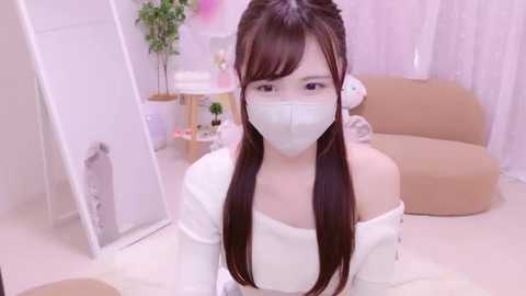 Media: Video of an East Asian woman with long brown hair, wearing a white off-shoulder top and face mask, sitting in a modern, pastel-colored room with beige furniture and plants.