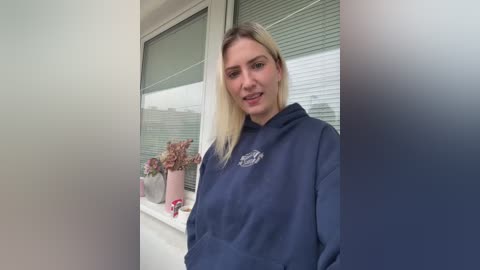 Media: Video of a fair-skinned woman with straight, blonde hair, wearing a dark blue hoodie, standing by a window with a potted plant and blinds, smiling slightly.