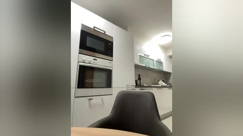 Media: A video of a modern, minimalist kitchen with white cabinets, stainless steel appliances, a microwave, and a dark gray dining chair in the foreground.