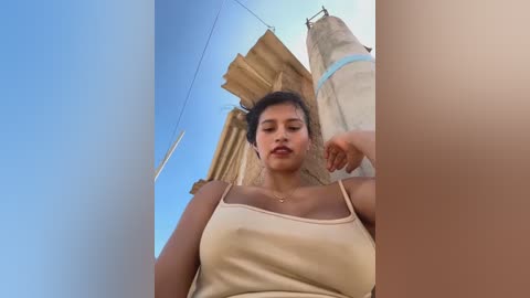 Media: A video of a young woman with medium brown skin and medium-length dark hair, wearing a beige tank top. She stands in front of a partially demolished building with a clear blue sky in the background.