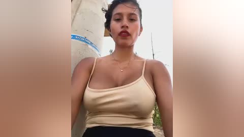 Media: Video of a Latina woman with medium skin tone, dark hair, and full lips, wearing a beige tank top and black pants, standing near a concrete pillar in a rural setting.