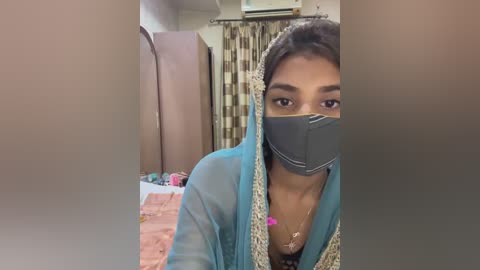 Media: Video of a young woman in a blue shalwar kameez, wearing a black face mask, standing indoors with beige curtains and a bed visible in the background.