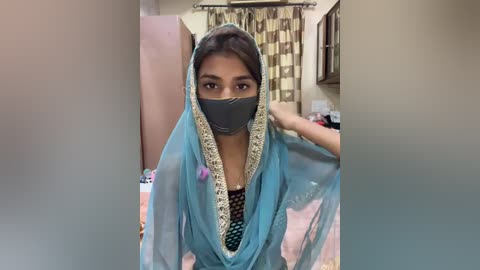 Media: Video of a woman in a light blue burqa with intricate embroidery, wearing a black face mask, in a modest Indian home with pink walls, a bed, and a window with beige curtains.