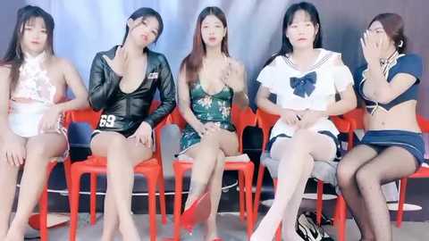 Media: Video of five East Asian women in revealing outfits, including a black leather top, a green floral dress, and a white sailor outfit, seated on orange plastic chairs against a white backdrop.