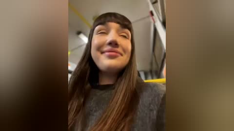 Media: A video of a young woman with long brown hair, wearing a gray robe, smiling widely. The background features a blurred, dimly lit room with white walls and a ladder.