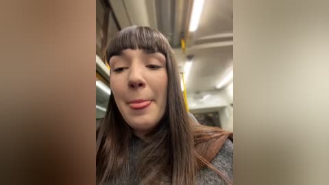 Media: A video of a young woman with straight brown hair and bangs, wearing a gray sweater, smiling inside a dimly lit subway car with fluorescent lights.