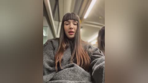 Media: Video of a young woman with long, straight, brown hair and bangs, wearing a gray sweater, standing in a subway car with fluorescent lighting and metal poles.