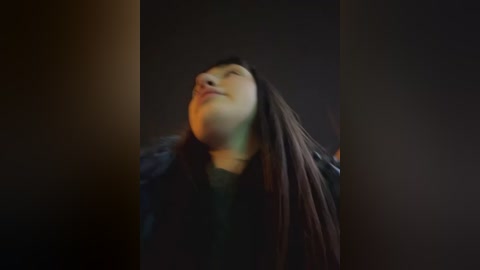 Media: A dimly lit video captures a young Asian woman with long, straight black hair, wearing a dark jacket, standing in a dimly lit room with a blurred background. Her eyes are closed, and she has a serene expression.