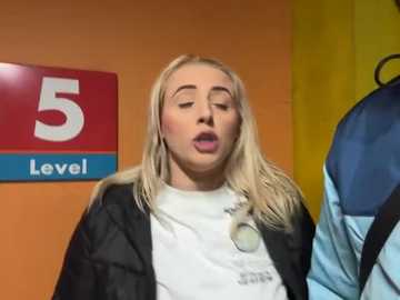 Media: Video of a blonde woman with long hair, wearing a black jacket and white shirt, mouth open in apparent surprise. Background shows a red and blue \"5 Level\" sign and a yellow wall.