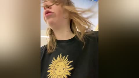 Media: A video of a young woman with fair skin, blonde hair, and closed eyes, wearing a black T-shirt with a sun graphic, captured in a blurry, grainy indoor setting.