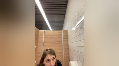 Media: Video of a modern, clean restroom with a wooden panel wall, white tiled floor, and a black ceiling with recessed lights. A young woman with long brown hair looks down, wearing a black top.