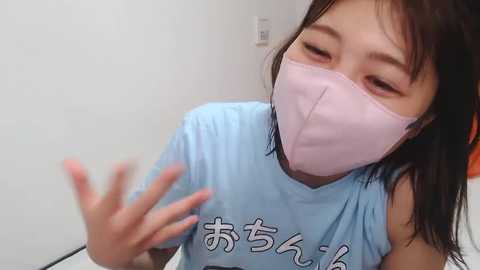 Media: Video of an Asian woman with straight black hair, wearing a light blue t-shirt and a pink surgical mask, making a hand gesture against a plain white wall.