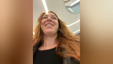 Media: A video of a smiling Caucasian woman with long, wavy red hair, wearing a black top, taken from a low angle, showing her from the neck up.