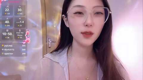 Media: Video of an Asian woman with fair skin, long dark hair, and clear glasses, wearing a white top, standing indoors with a blurred background.