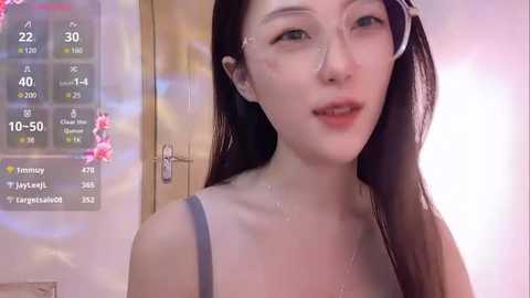 Media: Video of an Asian woman with long, wet black hair, wearing glasses, and a grey tank top, standing in a bathroom with a digital clock displaying 10:50, and a flowered background.