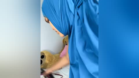 Media: Video of a person in blue medical scrubs, wearing a blue head covering, tending to a plush bear with a pink bow and black eyes on a white surface.