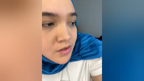 Media: Video of a young woman with light brown skin, wearing a blue hijab and white t-shirt, gazing introspectively, blurred background.