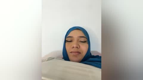 Media: Video of a serene young woman with a light brown complexion, wearing a blue hijab and a soft smile, lying on a white pillow against a plain white background.