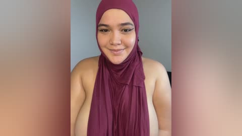 Media: Video of a light-skinned woman with a medium build, wearing a maroon hijab and a sleeveless top, smiling softly. Background is blurred, focusing on her.