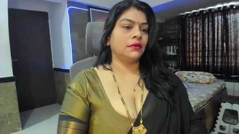 Media: Video of a South Asian woman with long black hair, wearing a gold blouse with a deep neckline and gold necklace, in a modern bedroom with beige walls, a bed, and dark curtains.