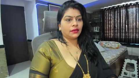 Media: Video of a South Asian woman with medium skin tone, long black hair, wearing a gold and black sari, sitting in a modern bedroom with a bed, curtains, and decorative items.