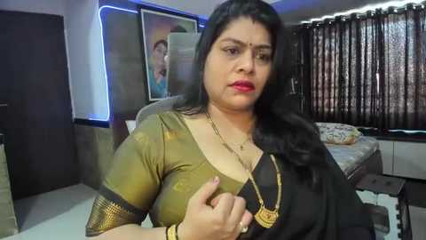 Media: Video of a middle-aged South Asian woman with long black hair, wearing a gold blouse and black sari, standing in a modern bedroom with a bed, curtains, and framed photos on the wall.